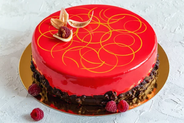 Mousse cake with red mirror glaze, chocolate decor and fresh raspberries on a textured white background.