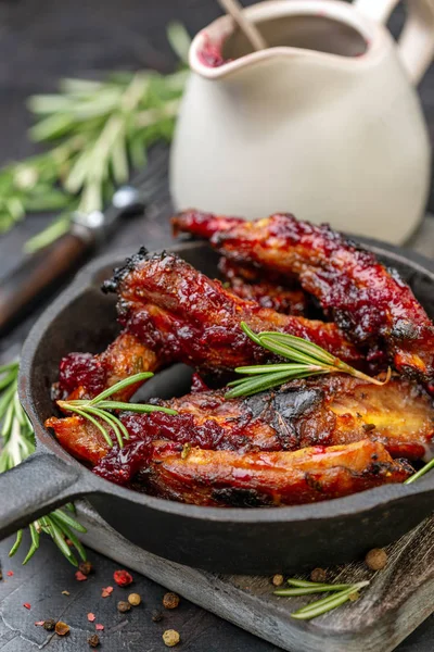 Cast iron frying pan with baked honey pork ribs and rosemary on wooden serving board, jug with cherry sauce on textured dark background, selective focus.