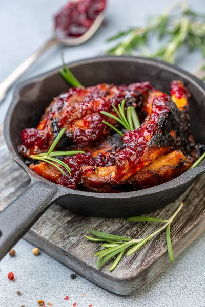 Cast iron pan with baked pork ribs, rosemary, spoon with cherry sauce on wooden serving board, selective focus.