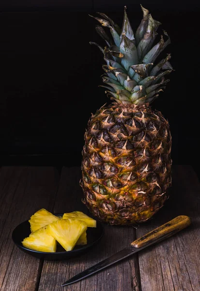 Sliced pineapple on old rustic desk with knife and with whole pineapple behind. Black background.