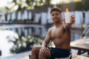 Güneş güvenlikli uygulamaları tanıtan genç bir adam, açık hava faaliyetleri sırasında cilt bakımının önemini vurgulayarak havuz kenarında güneş kremi sürüyor.