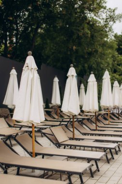 Boş güneş şezlongları ve kapalı beyaz şemsiyeler bir yüzme havuzunun yanında duruyor ve misafirler için sakin bir beklenti ortamı yaratıyor.