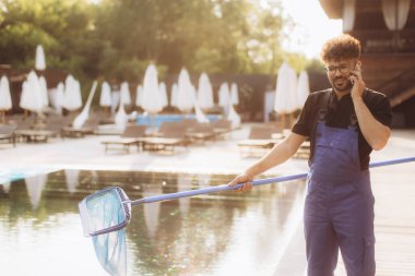 Güneşli bir yaz gününde havuz temizleyicisi mavi tulum giyip telefonla konuşuyor.