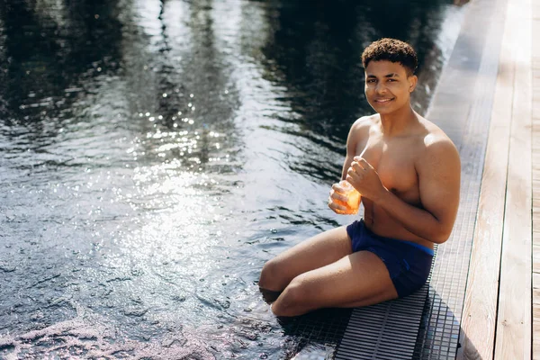 Yüzme havuzunda uzanan genç adam, güneşli bir yaz tatilinde güneşlenirken ferahlatıcı bir portakal kokteylinin tadını çıkarıyor.