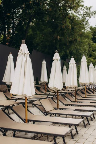 Boş güneş şezlongları ve kapalı beyaz şemsiyeler bir yüzme havuzunun yanında duruyor ve misafirler için sakin bir beklenti ortamı yaratıyor.