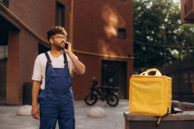Yemek teslimatı için termal bir sırt çantası tutarken akıllı telefon kullanan kurye hareketli şehir sokaklarında geziniyor.