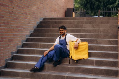 Teslimatçı ara veriyor. Yanında sarı termal çantasıyla merdivenlerde oturuyor.