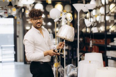 Işıkçı dükkanında lamba seçen gözlüklü genç bir adam. O, raflarda sergilenen çeşitli ışıklandırma ürünleriyle çevrili bir masa lambasını tutarken gülümsüyor..