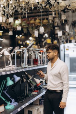 Beyaz gömlekli bir adam çağdaş bir aydınlatma mağazasında masa lambalarını inceliyor. Çeşitli avize modelleri, evler ve ofisler için geniş bir şık ışıklandırma seçeneklerini göstermektedir.