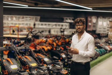 Genç adam bir hırdavatçı koridorunda çim biçme makinelerinin ortasında duruyor, modern aletler ve ekipmanlarla çevrili iken seçenekleri gözden geçiriyor..