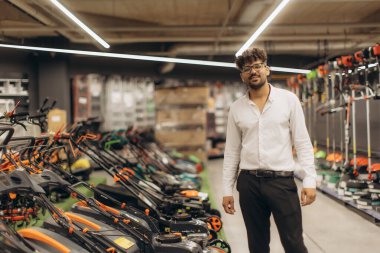 Bahçe ekipmanları ve aletlerle çevrili bir hırdavatçıda poz veren kendine güvenen bir mağaza çalışanı. Parlak ışıklandırma ve organize düzen profesyonel ve karşılayıcı bir perakende atmosfer yaratıyor..