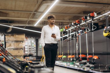 Perakende satış reyonundaki bir adam elinde akıllı telefon tutarken çeşitli ekipman ve makine seçeneklerini araştırıyor..