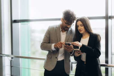 İki iş adamı, modern ofis ortamında akıllı telefonları kullanarak, teknoloji, iletişim ve takım çalışmalarını vurgulayarak, ortak bir tartışmada fikir ve bilgi paylaşımı yapıyorlar..