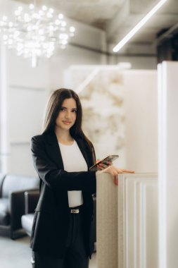 Çağdaş bir ofiste elinde akıllı bir telefonla resmi giyinmiş profesyonel bir kadın. Modern iş kültürü ve işyeri dinamiklerini yansıtan güven ve profesyonellik sergiliyor..