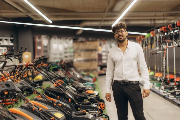 Bahçe ekipmanları ve aletlerle çevrili bir hırdavatçıda poz veren kendine güvenen bir mağaza çalışanı. Parlak ışıklandırma ve organize düzen profesyonel ve karşılayıcı bir perakende atmosfer yaratıyor..