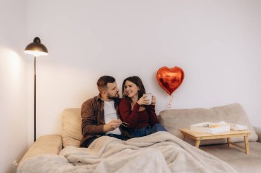 Kanepede sıcak bir anın tadını çıkaran bir çift ellerinde kupalar ve kırmızı bir kalp balonu olan..