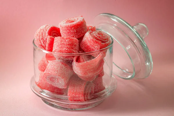 Chewy sweets and strawberry and cherry flavoured gummy candy concept with close up on sweet and sour red sour belts covered in sugar in a clear glass jar isolated on a pink background