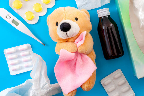 Cold and flu treatment and pediatrics medicine concept with a teddy bear holding a blanket surrounded by  a thermometer, bottle of cough syrup, sore throat lozenges, pills, capsules and paper tissue