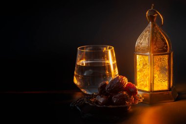 Müslüman dini geleneği, kutsal Ramazan ayı, İslam ve iftar konsepti temalı bir kase hurma, dua boncukları, bir bardak su ve siyah arkaplan üzerine Arapça fener.