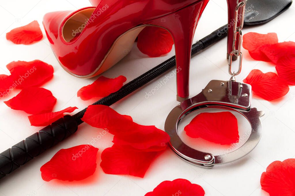 Kinky sex and BDSM games concept with a pair of red and shiny high heel stilettos, a leather crop under the shoes, a pair of metal handcuffs and rose petals surrounding the scene