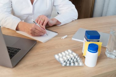 Stresli bir kadın ofis masasında, bir elinde ilaçlarını, diğer elinde de bir bardak su tutuyor.