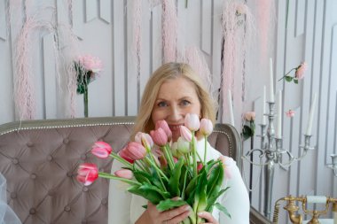 Zarif bir kadın şık, güzel dekore edilmiş bir yerde dururken canlı pembe laleler tutuyor.