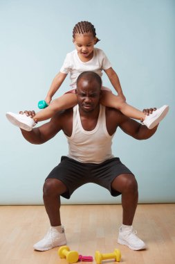 Handsome black young father is doing squats with his cute little daughter riding him. He is having trouble with additional weight, which is funny to her.