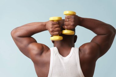 Black bold man in sleeveless white shirt doing overhead tricep extention with two light yellow-collored plastic dumbbells from behind. pale blue background. Tensing biceps. Fitness workout.