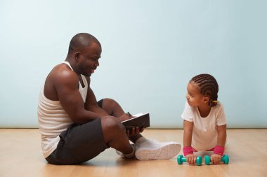Yakışıklı siyah genç baba nasıl halter ile egzersiz için onun sevimli küçük kızı talimat, notlar yapma. Soluk mavi arka plana karşı. İtme ler. Çok dikkat ediyor..