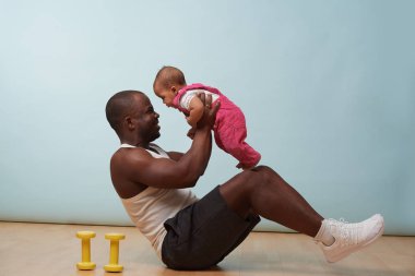Handsome black young father is doing basic crunches while holding his tiny little baby daughter in hands. He is having trouble with additional weight.