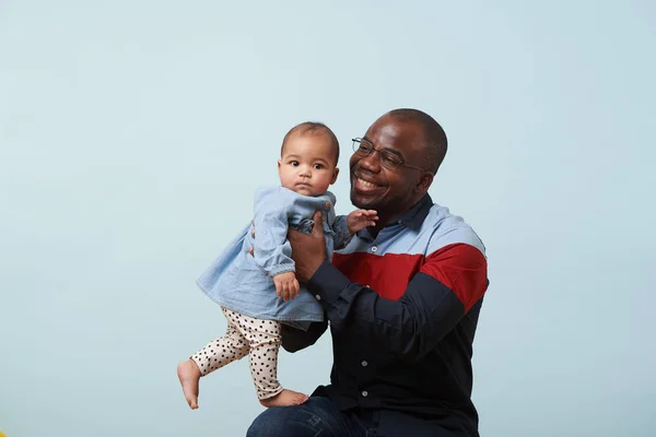 Happy father in big stripe shirt holds his little tiny baby daughter in arms against pale blue background. Looking at her with love. bare feet