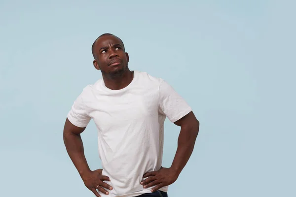 Handsome serious bold black man in white T-shirt with hands on waist, against pale blue background. Displeased, grumpy, unsatisfied, upset, angry.