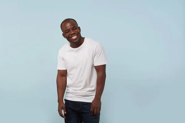 Handsome bold black man in white T-shirt grimacing against pale blue background. Relaxed, arms down, giggling, eyes closed. Laughing at something funny