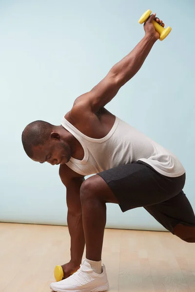 Handsome black bold man in sleeveless white shirt doing waist rotations with two light yellow-collored plastic dumbbells. pale blue background. Fitness workout. Touching floor. Stretching.