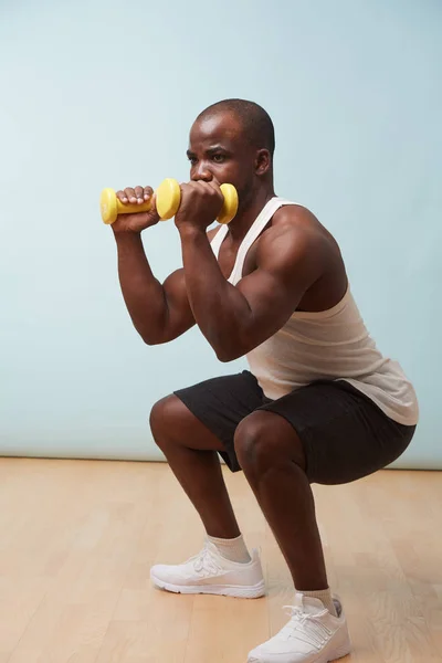 Handsome black bold man in sleeveless white shirt doing squats with two light yellow-collored plastic dumbbells. pale blue background. Fitness workout.