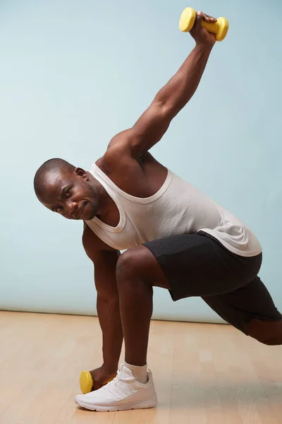 Handsome black bold man in sleeveless white shirt doing waist rotations with two light yellow-collored plastic dumbbells. pale blue background. Fitness workout. Touching floor. Stretching.