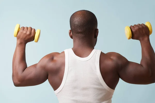 Black bold man in sleeveless white shirt lifting up two light yellow-collored plastic dumbbells from behind. pale blue background. Tensing biceps. Fitness workout.