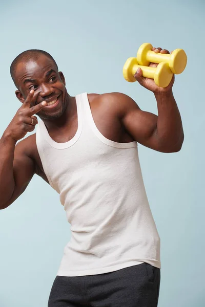 Handsome black bold man in sleeveless white is cheering up, pleased with him reaching the end of workout. pale blue background. Fitness workout. Victorious gestures. V sign.