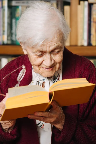 Porträtt av glada grå haired kvinna läser bok nära bokhylla hemma — Stockfoto