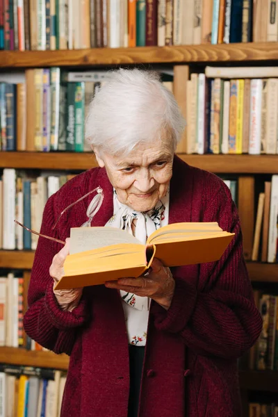 Porträtt av glada grå haired kvinna läser bok nära bokhylla hemma — Stockfoto