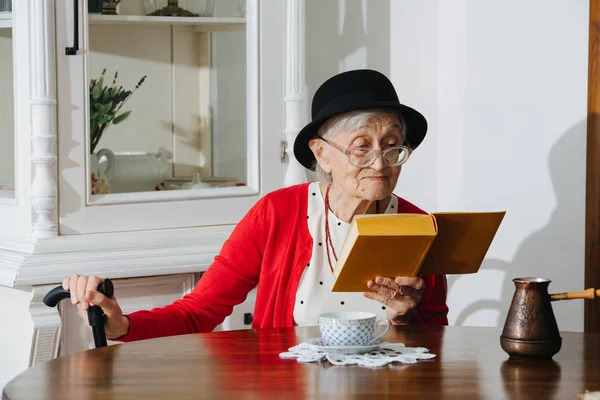 Kind grandmother is reading her book at dinner table at home. — Stock Photo, Image