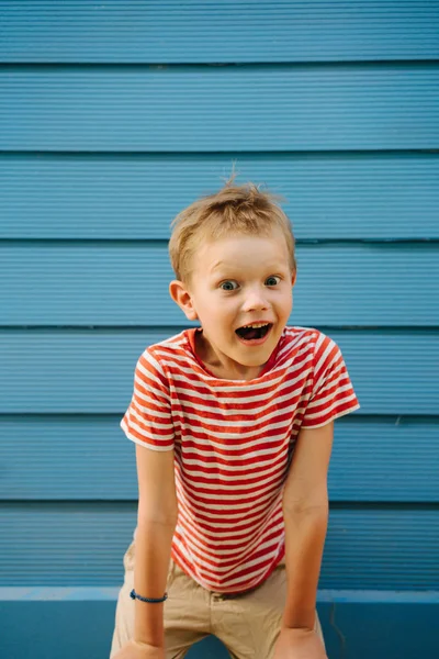 Cute little boy in striped T-shirt posing in front of blue house wall. — Stock Photo, Image
