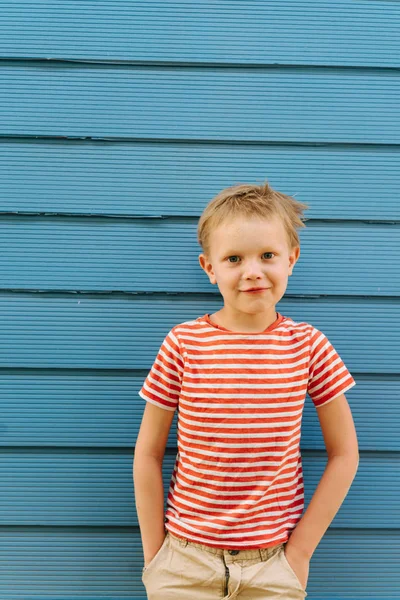 Cute little boy in striped T-shirt posing in front of blue house wall. — Stock Photo, Image