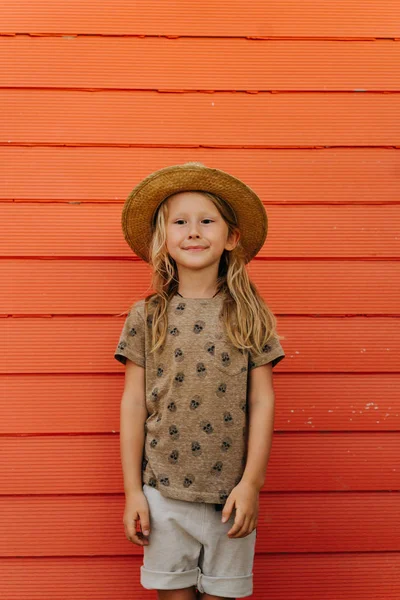 Little boy with long blond hair in summer hat posing in front of house wall. — Stock Photo, Image