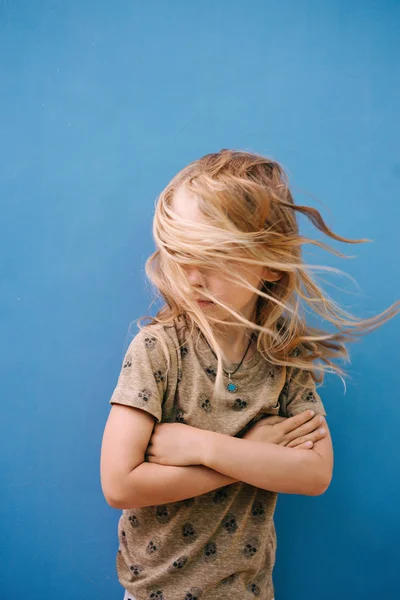 Niño con el pelo largo y rubio en camiseta blanca posando frente a la pared de la casa . — Foto de Stock