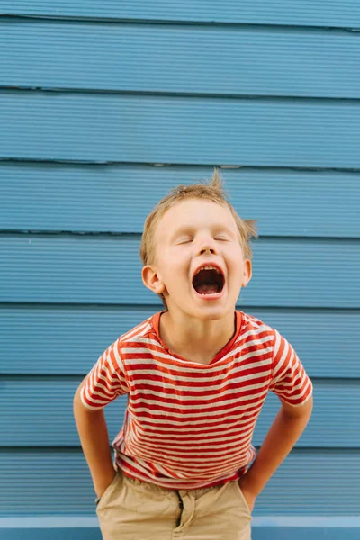 Lindo niño en camiseta rayada posando frente a la pared azul de la casa . — Foto de Stock