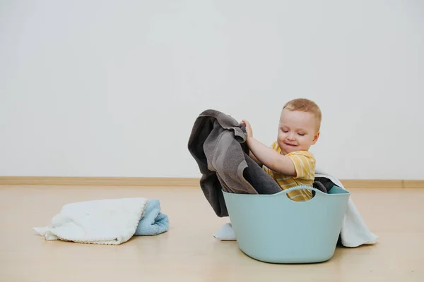 Happy toddler son sitting in laundry basin throwing towels aside — Stock Photo, Image