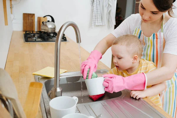 Joven mujer feliz en una cocina está lavando tazas y platos. Su hijito ayuda —  Fotos de Stock