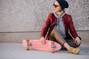 Genç, mutlu, açık tenli, esmer kadın kaykayıyla sokak zemininde oturuyor.