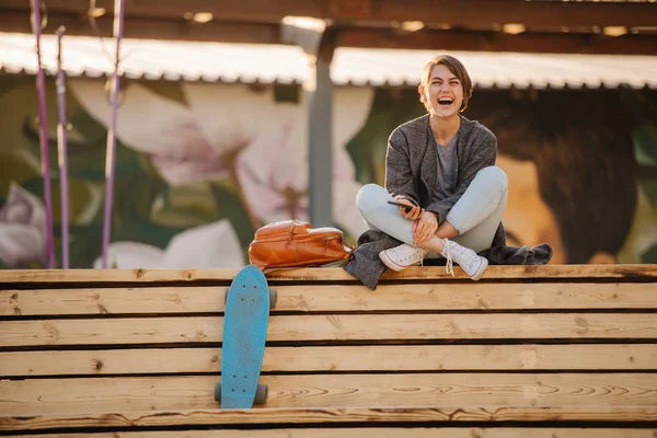 Mladá smějící se žena s skateboardem sedí na dvoustupňové lavičce. — Stock fotografie